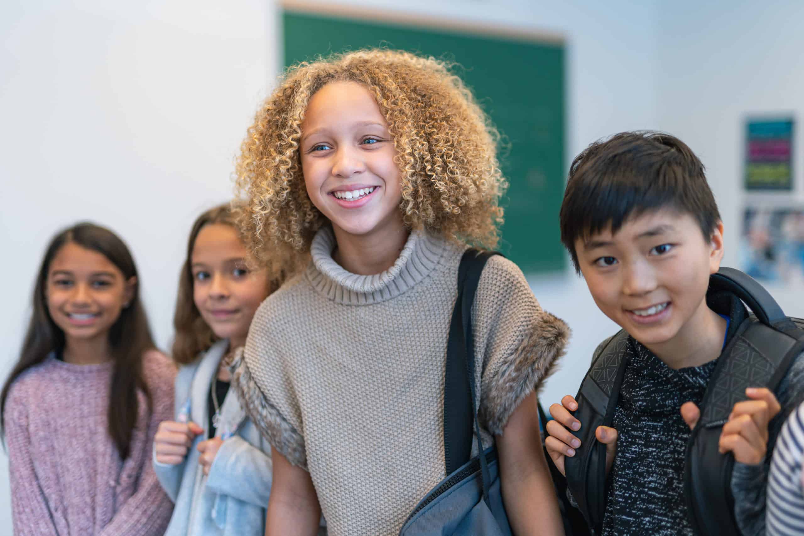A multi-ethnic group of elementary age students is standing in a row in their classroom. They are smiling and wearing their backpacks. The image is focused on a multi-ethnic girl at the center of the group. She is taller than her classmates.