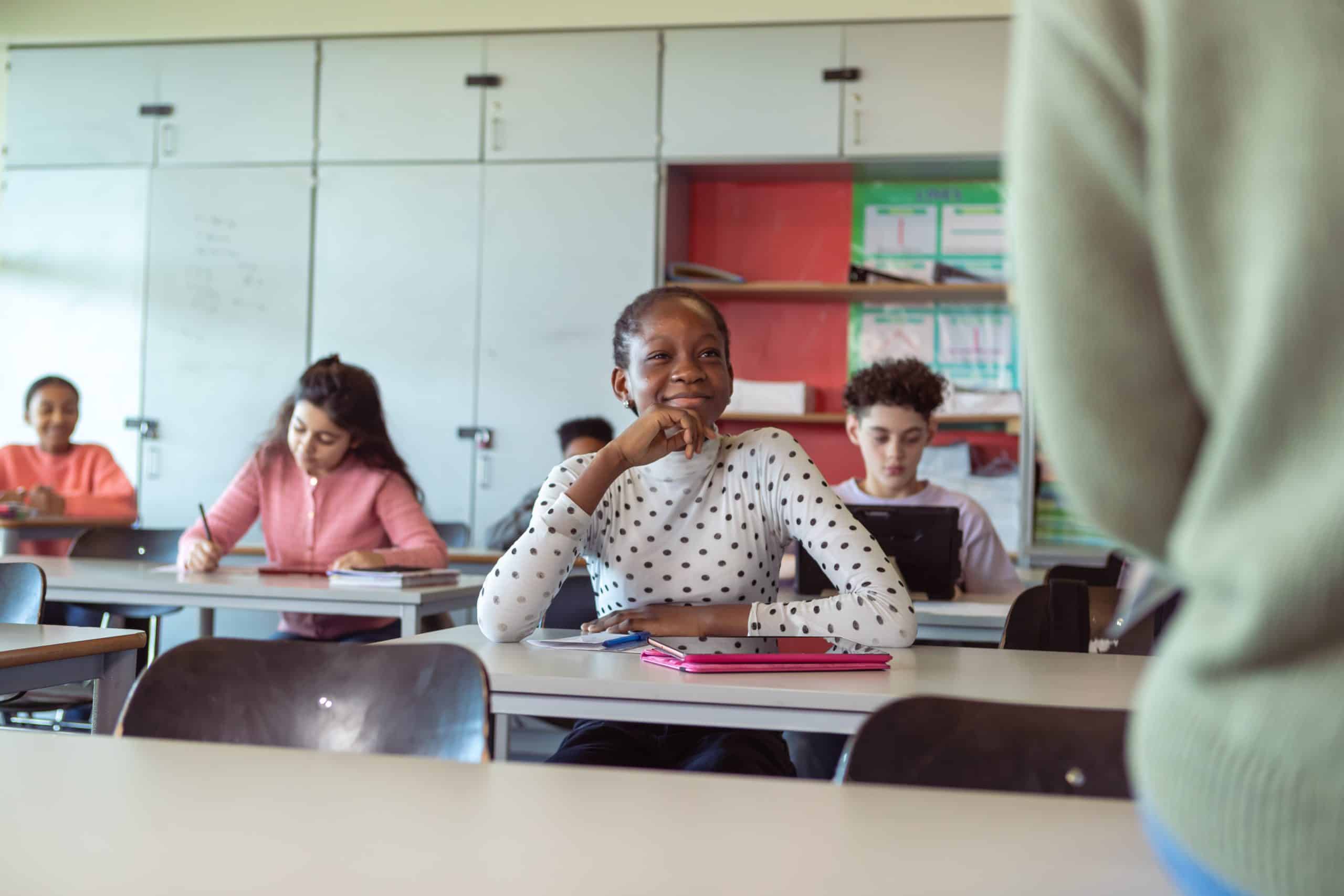 A multiracial group of students in elementary school sitting at desks take notes and listen as their teacher gives instructions during a class lecture. Selective focus on a cheerful black female students sitting at the front of the classroom.