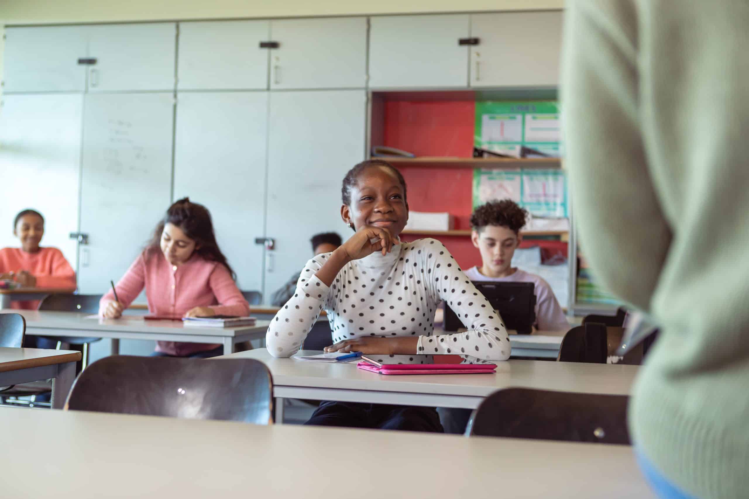 Attentive adolescent students listening to lecture in class A multiracial group of students in elementary school sitting at desks take notes and listen as their teacher gives instructions during a class lecture. Selective focus on a cheerful black female students sitting at the front of the classroom.
